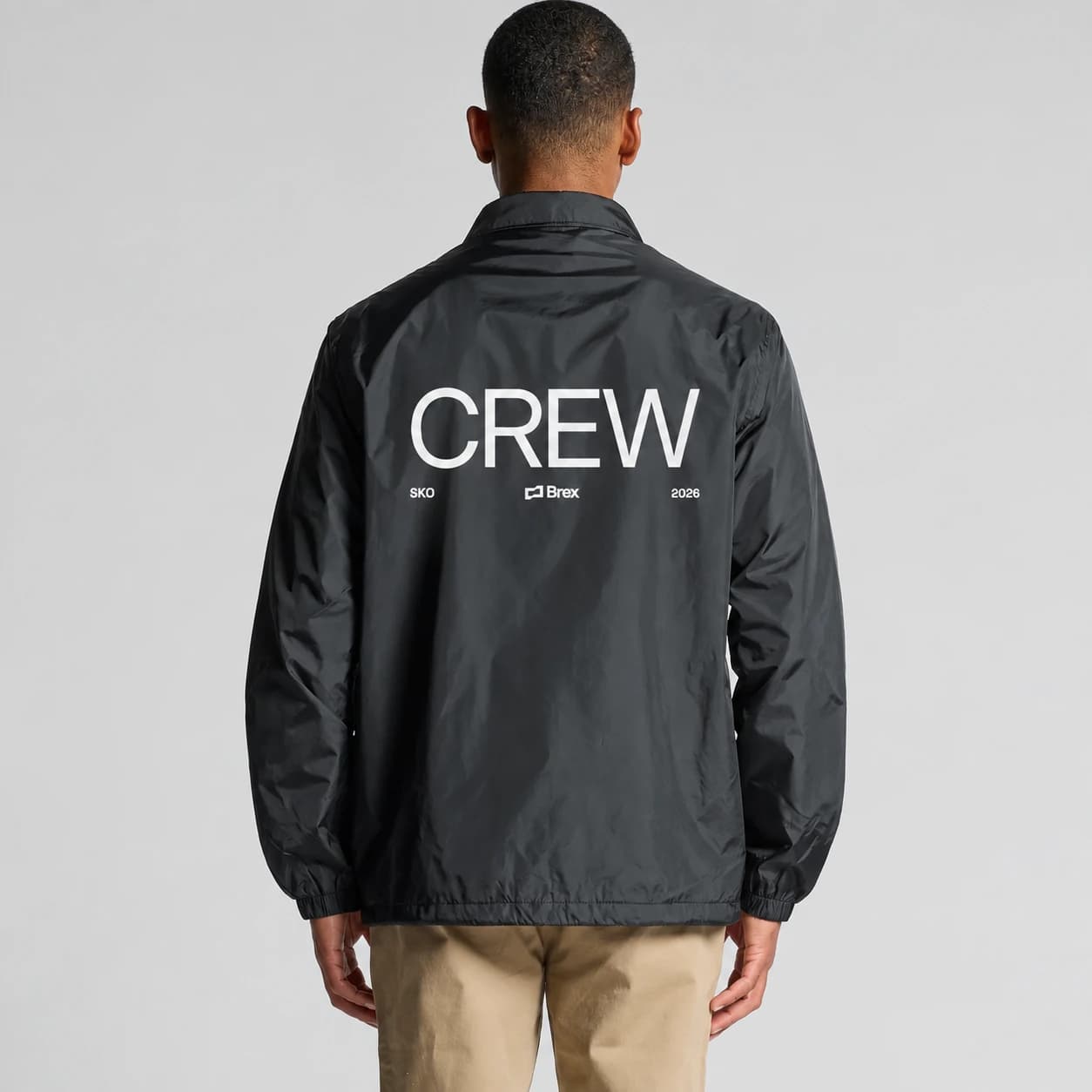 Studio image of a person wearing a black Brex coach jacket with white CREW, SKO and 2026 artwork across the back.
